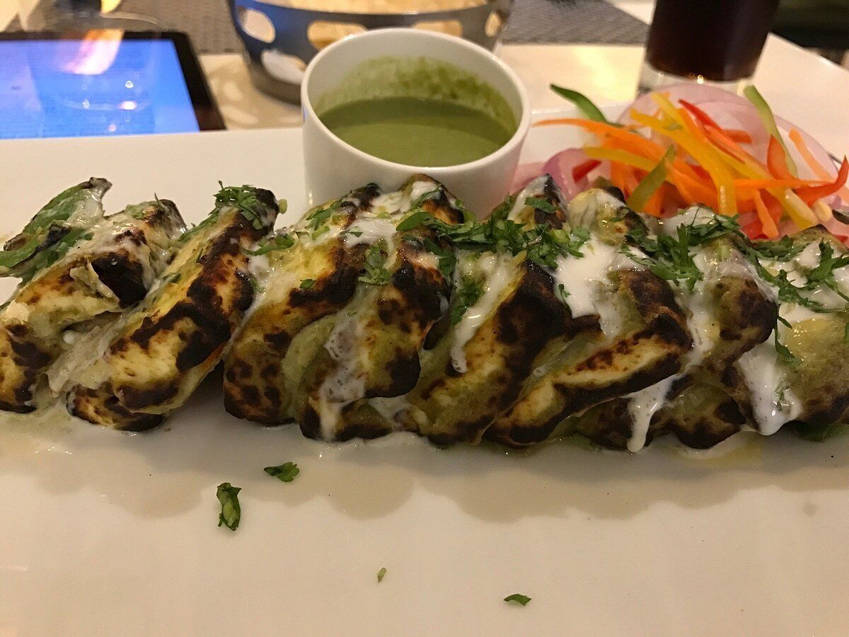 A plate of grilled chicken wings with a creamy sauce, a side of sliced vegetables, and a green sauce in a small bowl.