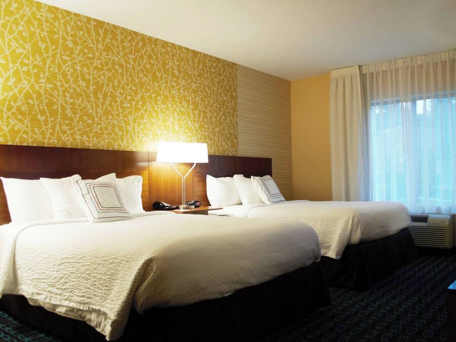 A hotel room with two beds, a bedside table with a lamp, a chair, and a window with curtains.