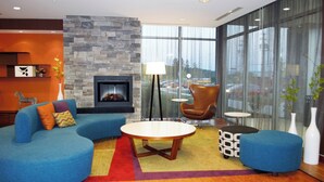 A modern living room with a stone fireplace, blue sofas, a round coffee table, and large windows offering a view of the parking lot. - Fairfield Inn & Suites Stroudsburg Bartonsville / Poconos (Stroudsburg)