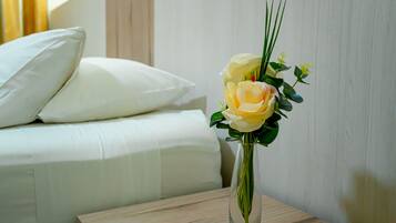 Una cama con ropa de cama y almohadas blancas, una mesita de noche con un florero transparente con flores, y una mesita auxiliar de madera.