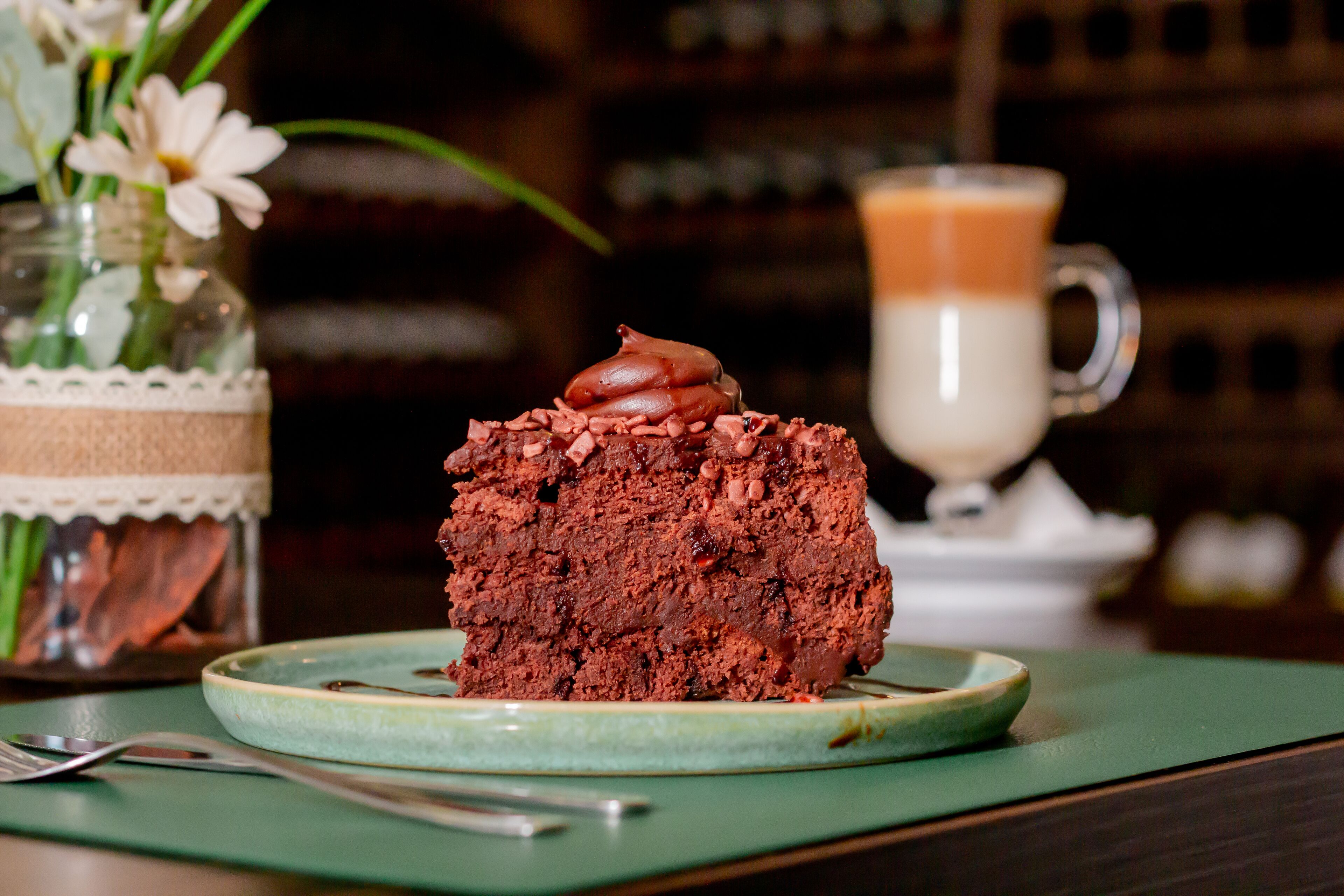 A slice of chocolate cake with whipped cream on a plate, a glass of coffee, and a jar of flowers in the background.