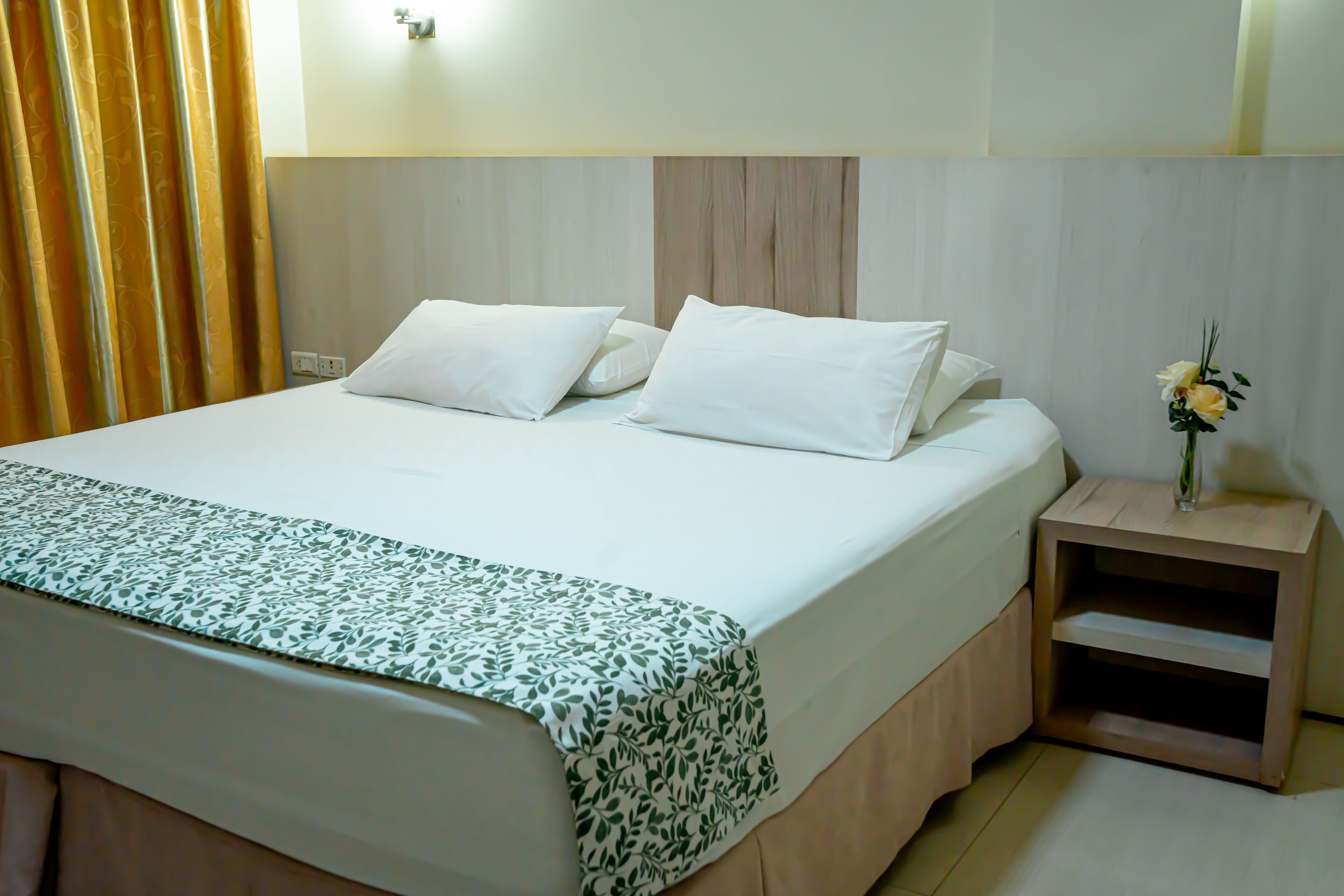 A neatly made bed with white linens and a green leaf-patterned throw, a bedside table with a vase of flowers, and a wooden headboard.