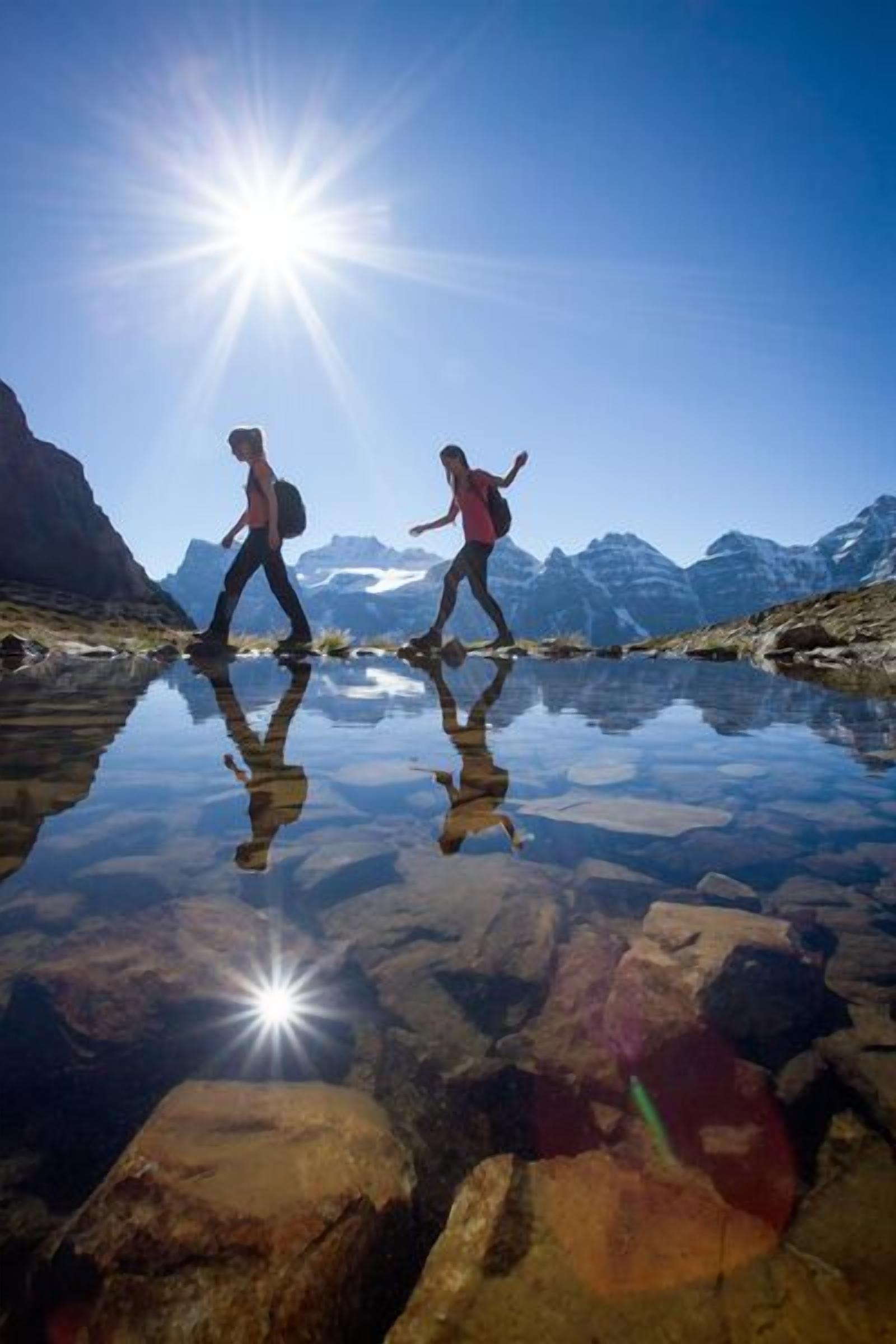 Dos personas caminando sobre una superficie acuática reflectante, con montañas al fondo y el sol brillando intensamente.