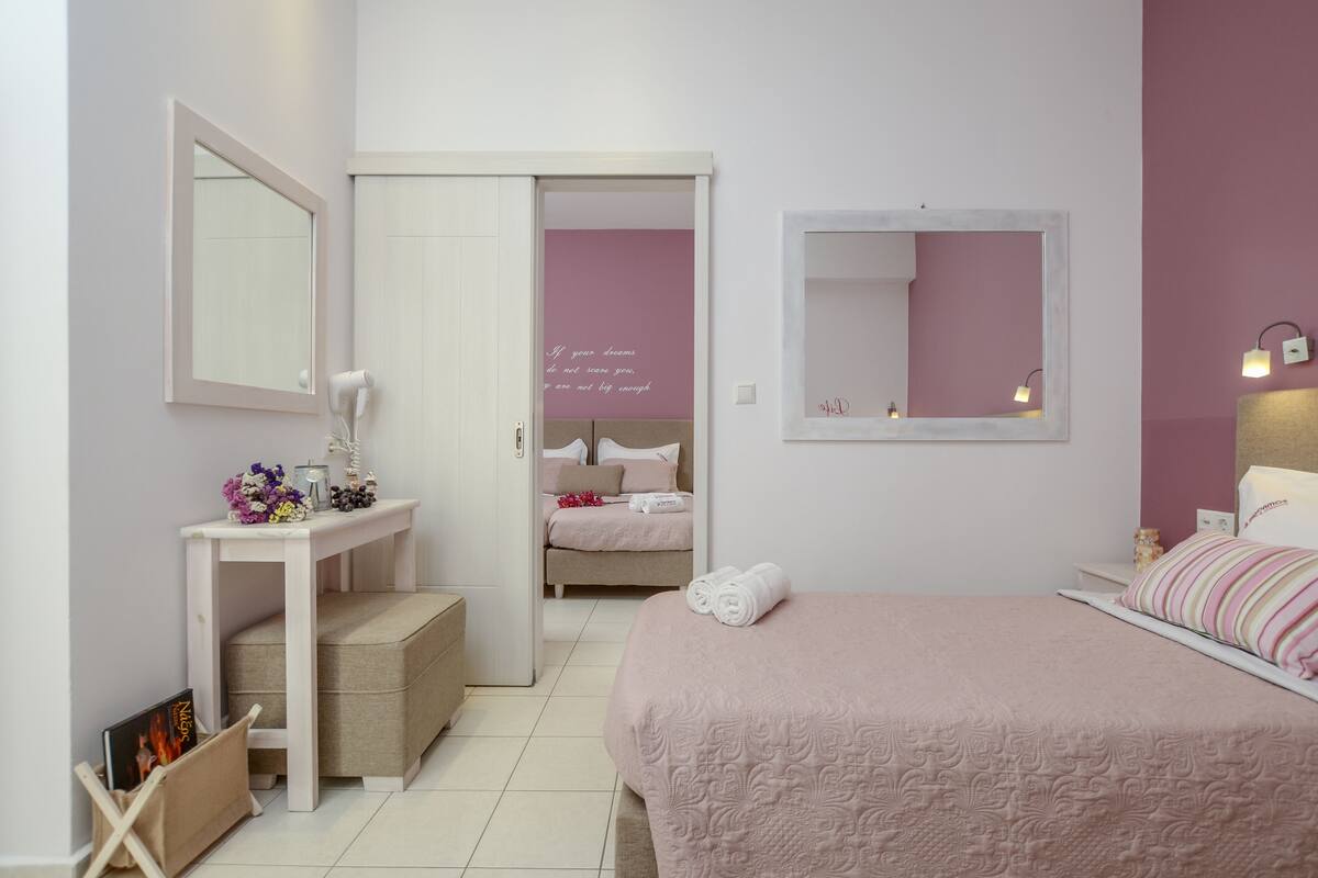 A bedroom with a bed, bedside table, and a mirror. There is a door leading to another room.