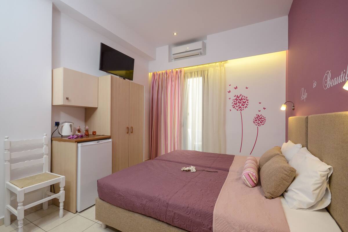 A bedroom with a bed, a chair, a small kitchenette, and a wall with a decorative tree and butterflies.