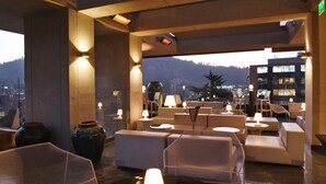 Un salón en la azotea con asientos y mesas, vistas a la ciudad y las montañas, y un jarrón grande.