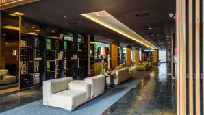 Un moderno hall de hotel con un mostrador de recepción alargado, una zona de estar y una amplia estantería llena de libros y dispositivos electrónicos.