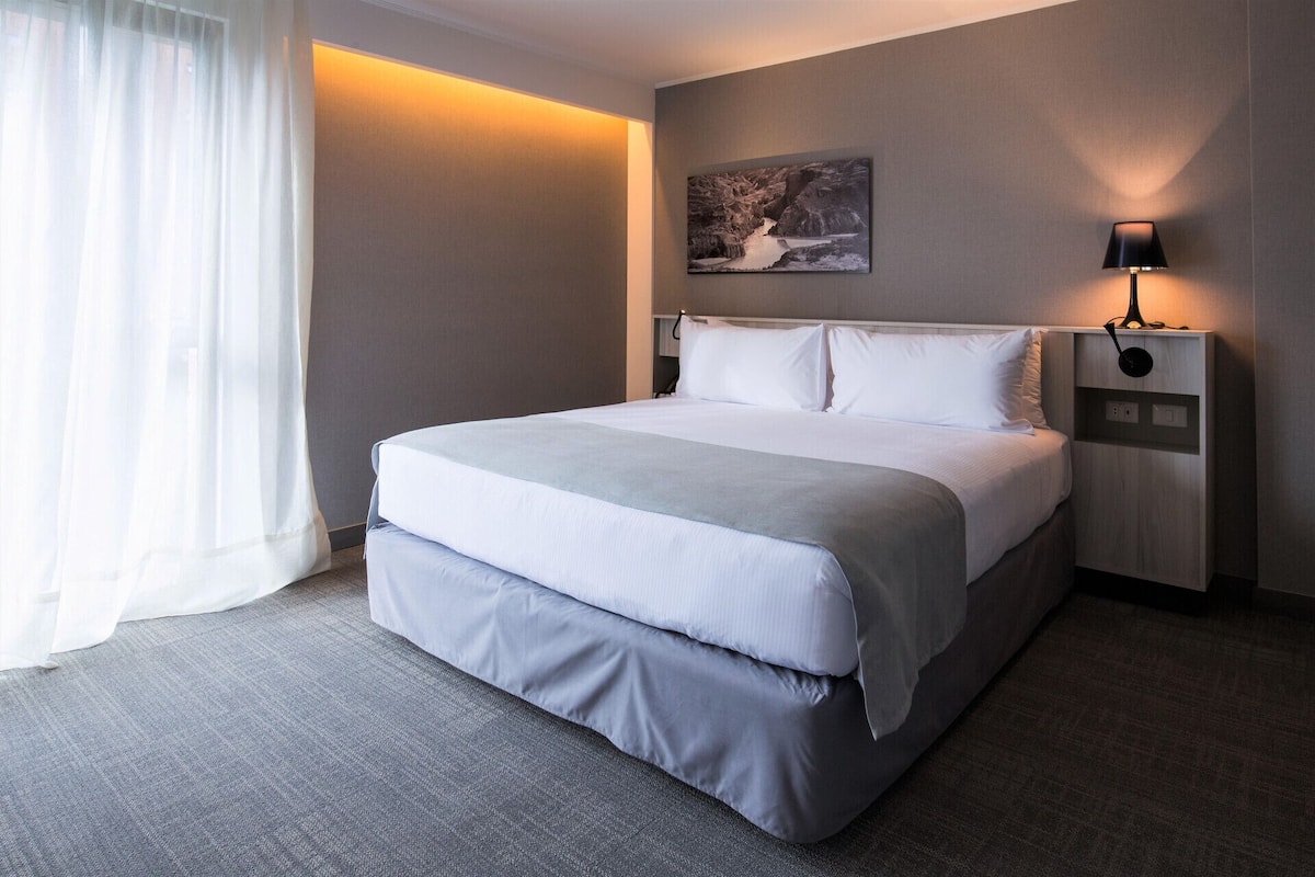 Une chambre d’hôtel avec un lit, une table de chevet, une lampe, un tableau au mur et une fenêtre avec des rideaux.