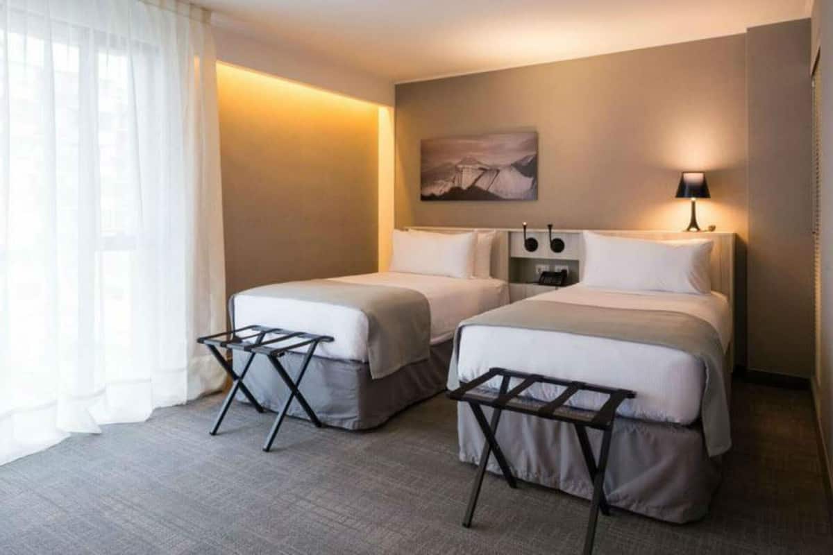 Une chambre d’hôtel avec deux lits, une fenêtre avec des rideaux légers, un tableau au mur et une lampe de chevet.