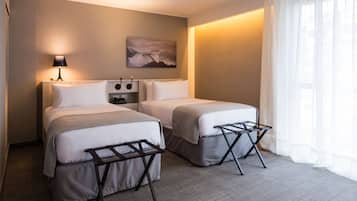 Habitación de hotel con dos camas, una mesita de noche, una lámpara, un cuadro y una ventana con cortinas traslúcidas.