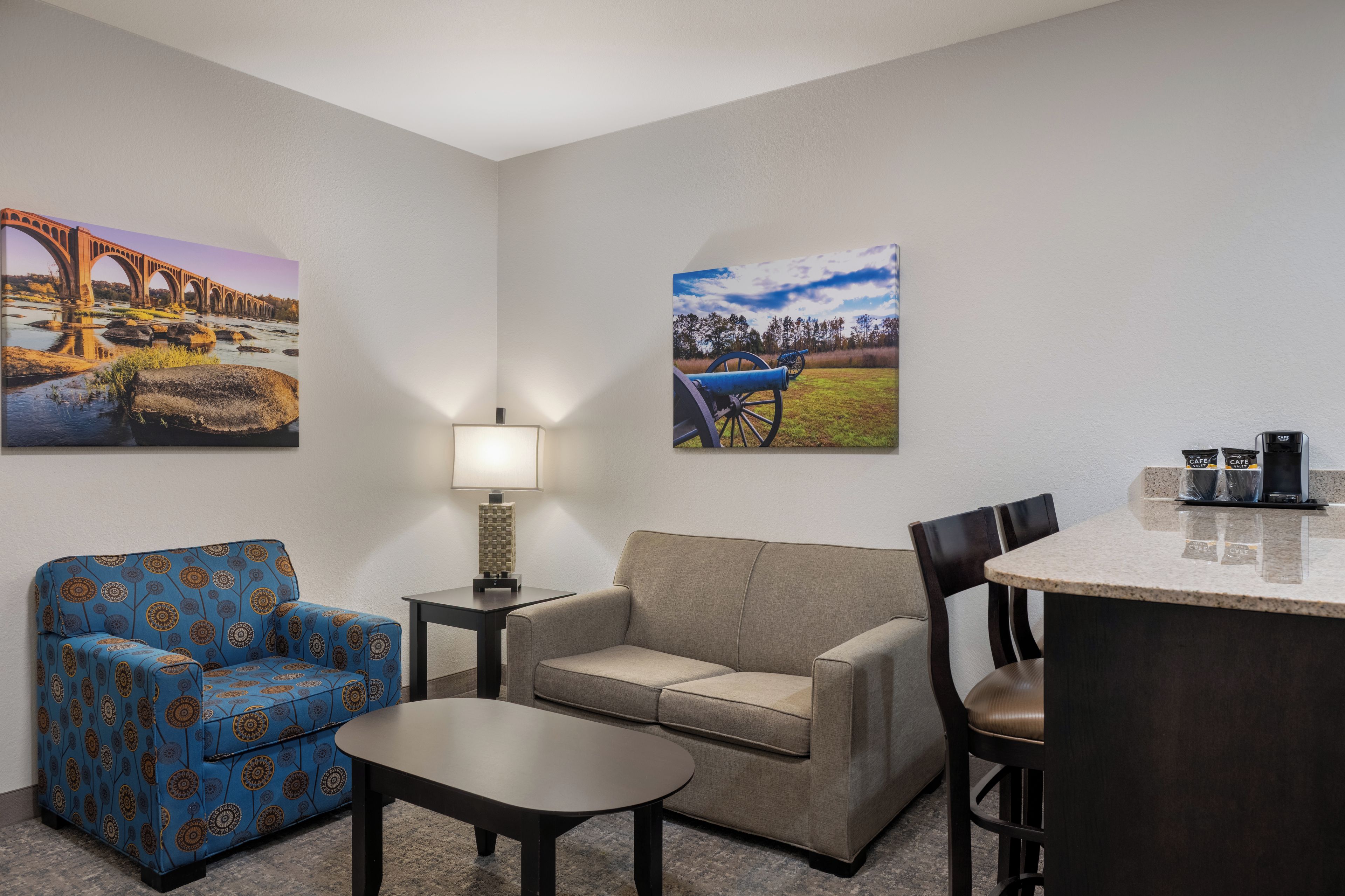 Habitación de hotel con sofá, sillón, mesa de centro y un pequeño comedor con mesa y sillas. Hay dos cuadros en las paredes.