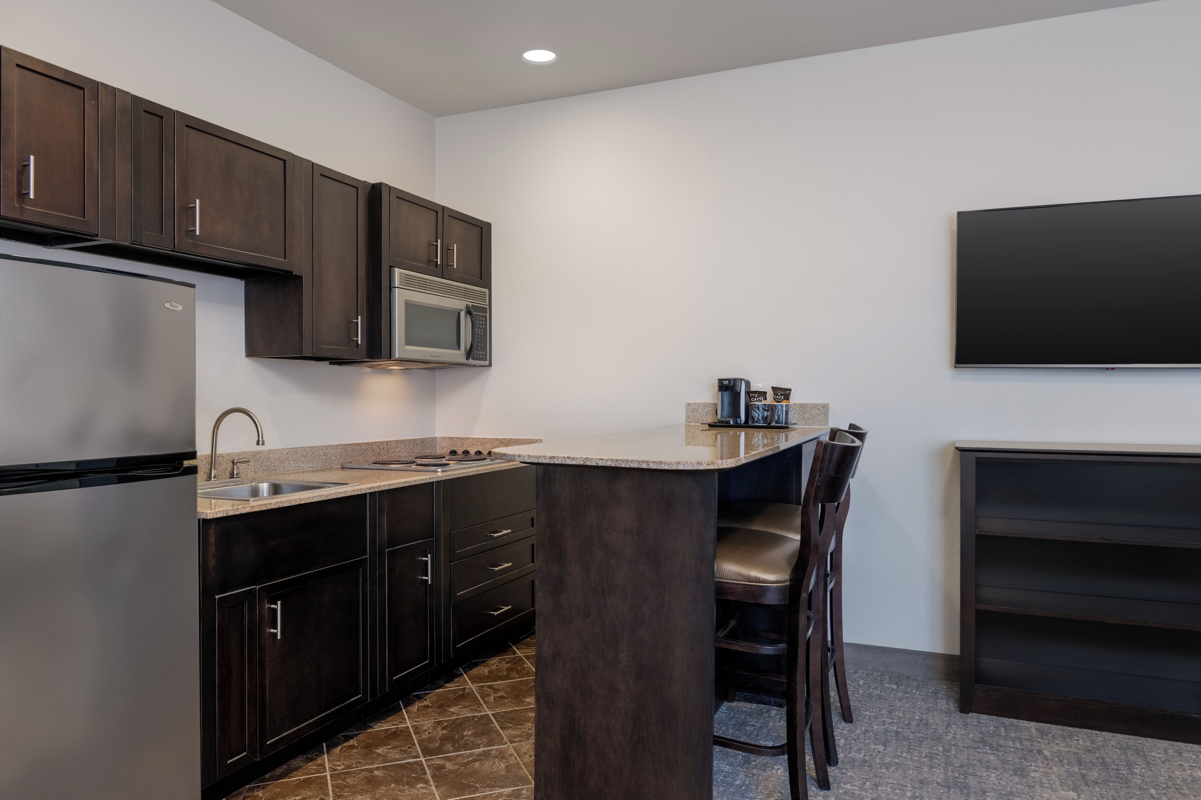 A modern kitchen with dark wood cabinets, a stainless steel refrigerator, a microwave, and a built-in island with bar stools.