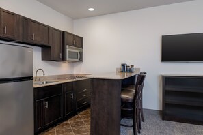 A modern kitchen with dark wood cabinets, a stainless steel refrigerator, a microwave, and a built-in island with bar stools. - Stay-Over Suites (Hopewell)