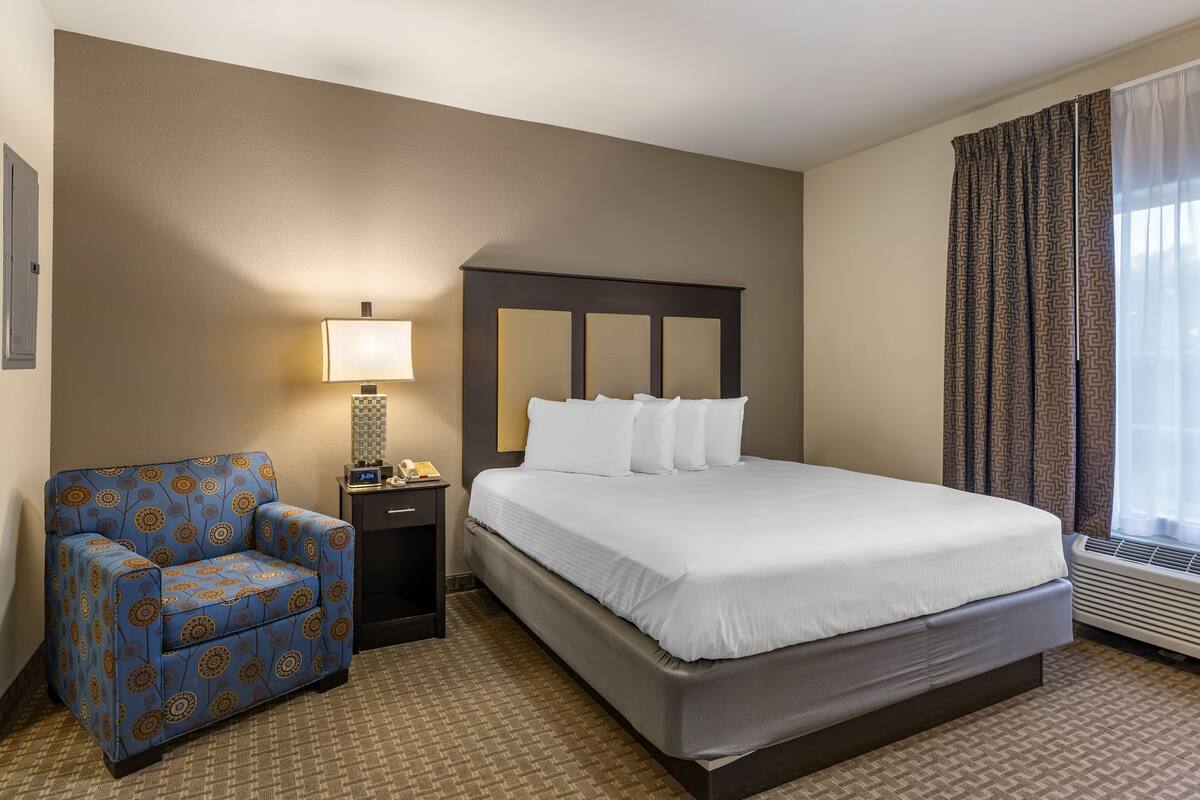 A hotel room with a bed, a chair, a nightstand, a lamp, and a window with curtains.