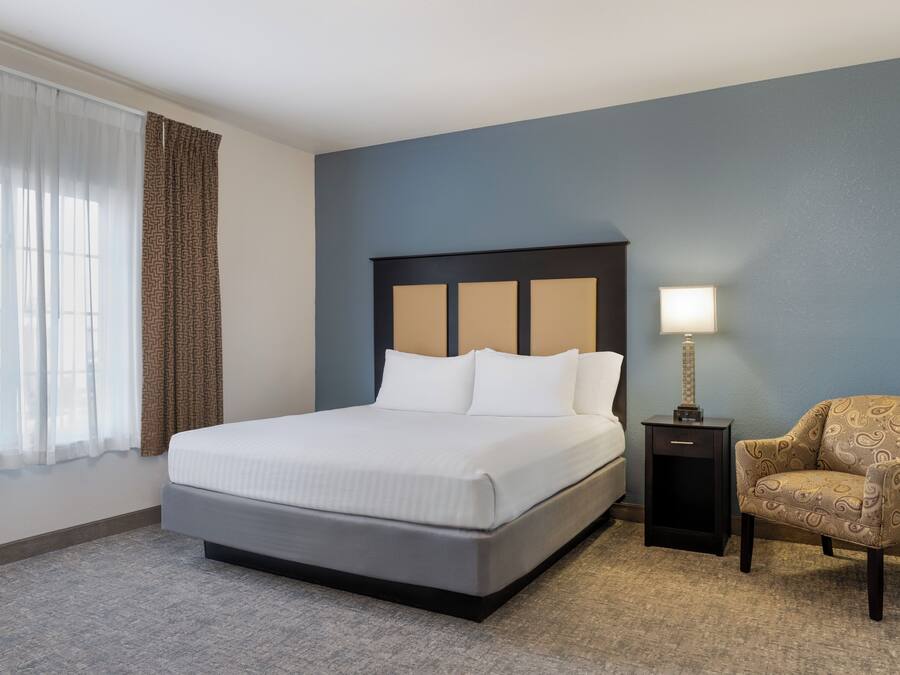 A hotel room with a large bed, a nightstand with a lamp, a chair, and a window with curtains.