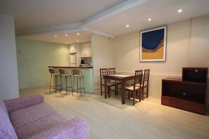A living room with a pink sofa, a dining table with four chairs, a kitchen area with a bar stool, and a large abstract painting on the wall.