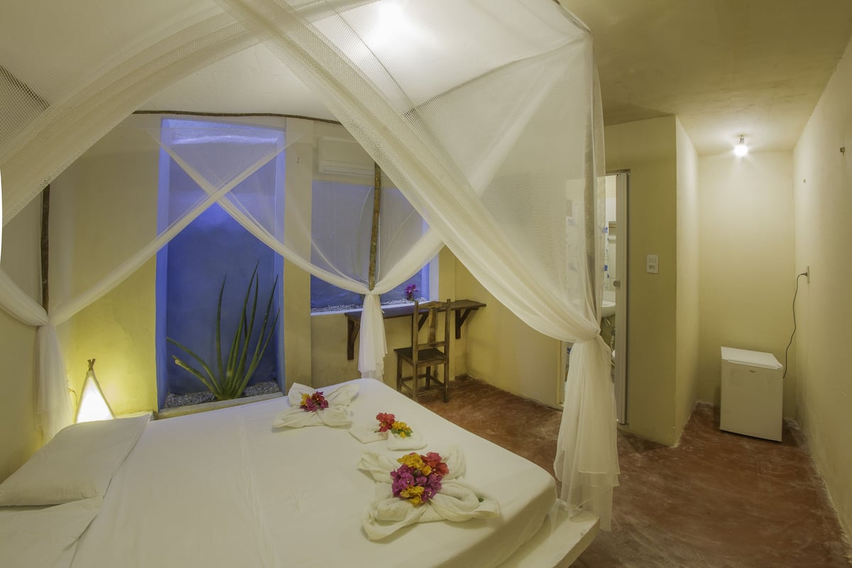 Ein Schlafzimmer mit Bett, Sessel, Nachttisch, Vorhängen an einem Fenster und einem kleinen Tisch mit Lampe.