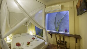 Ein Zimmer mit einem Bett unter einem Baldachin, einem kleinen Tisch, einem Stuhl, einem Fenster mit Blick auf Pflanzen und einer Klimaanlage.