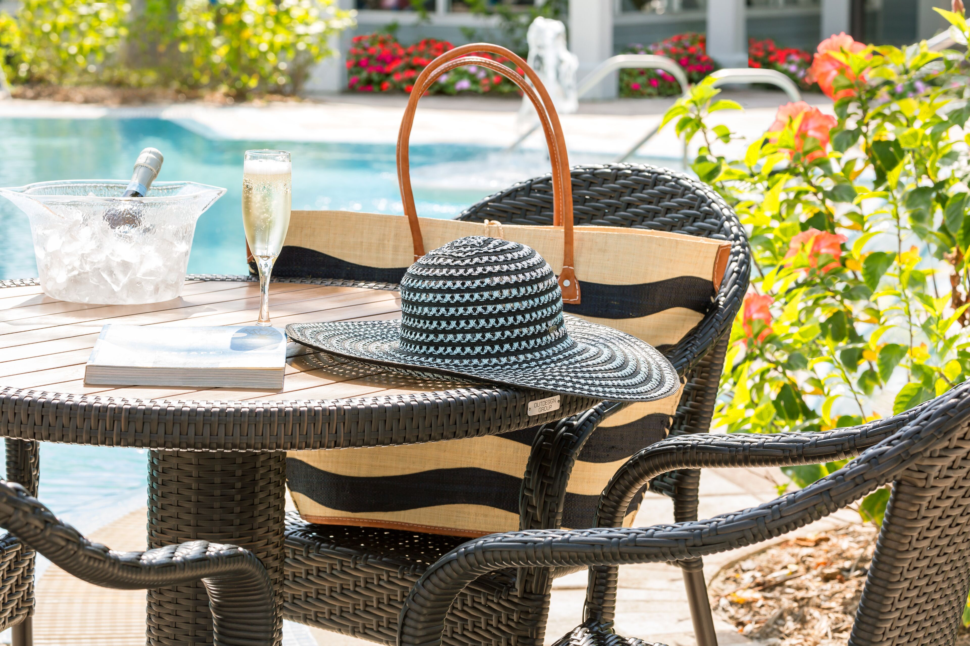 Une chaise en osier avec un sac à rayures et un chapeau, un verre de champagne et un seau à glace, le tout placé près d’une piscine.