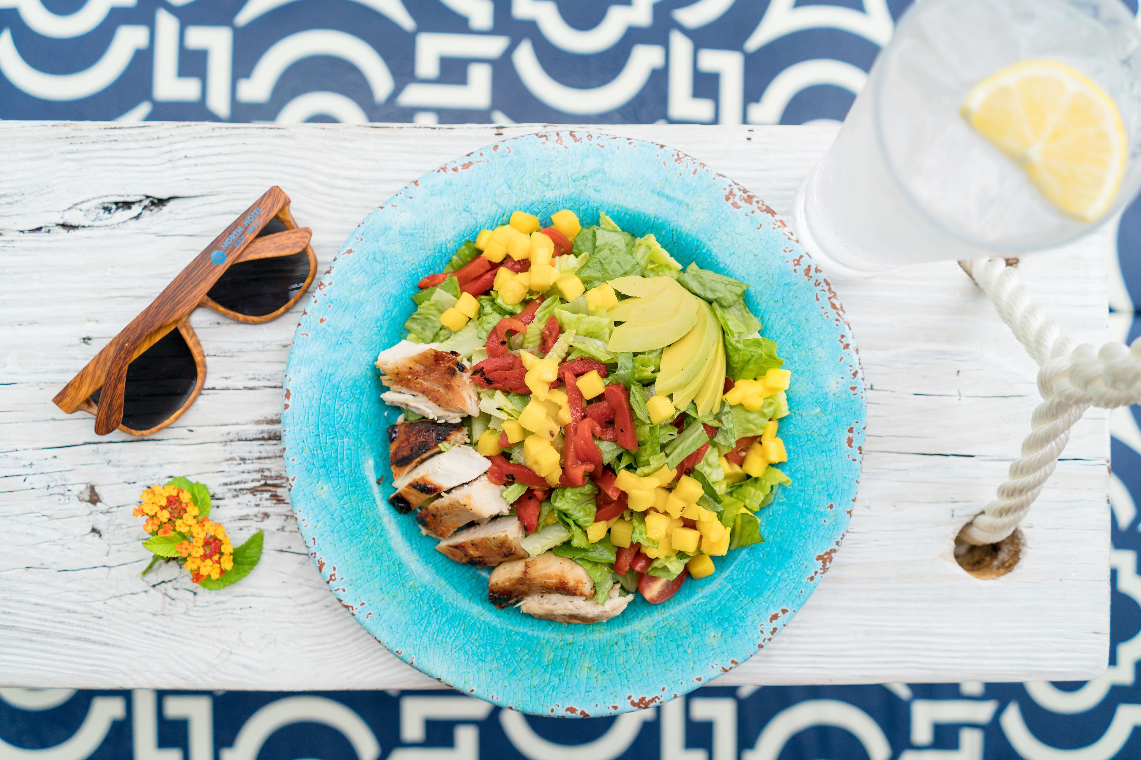 A blue plate with a salad, sunglasses, and a glass of water on a white wooden surface.