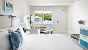 Ein modernes Hotelzimmer mit einem großen Bett, einer kleinen Sitzecke mit Tisch, einem Fenster mit Blick auf den Pool und einer Tür zum Badezimmer.