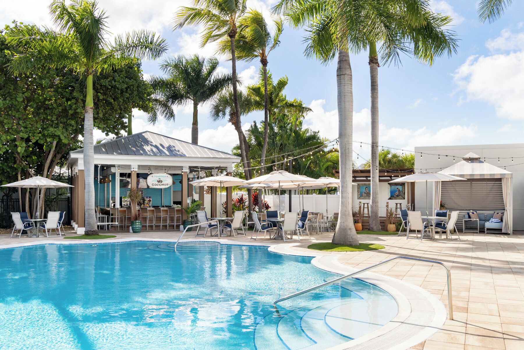 A pool area with lounge chairs and umbrellas, surrounded by palm trees and a building with a sign.