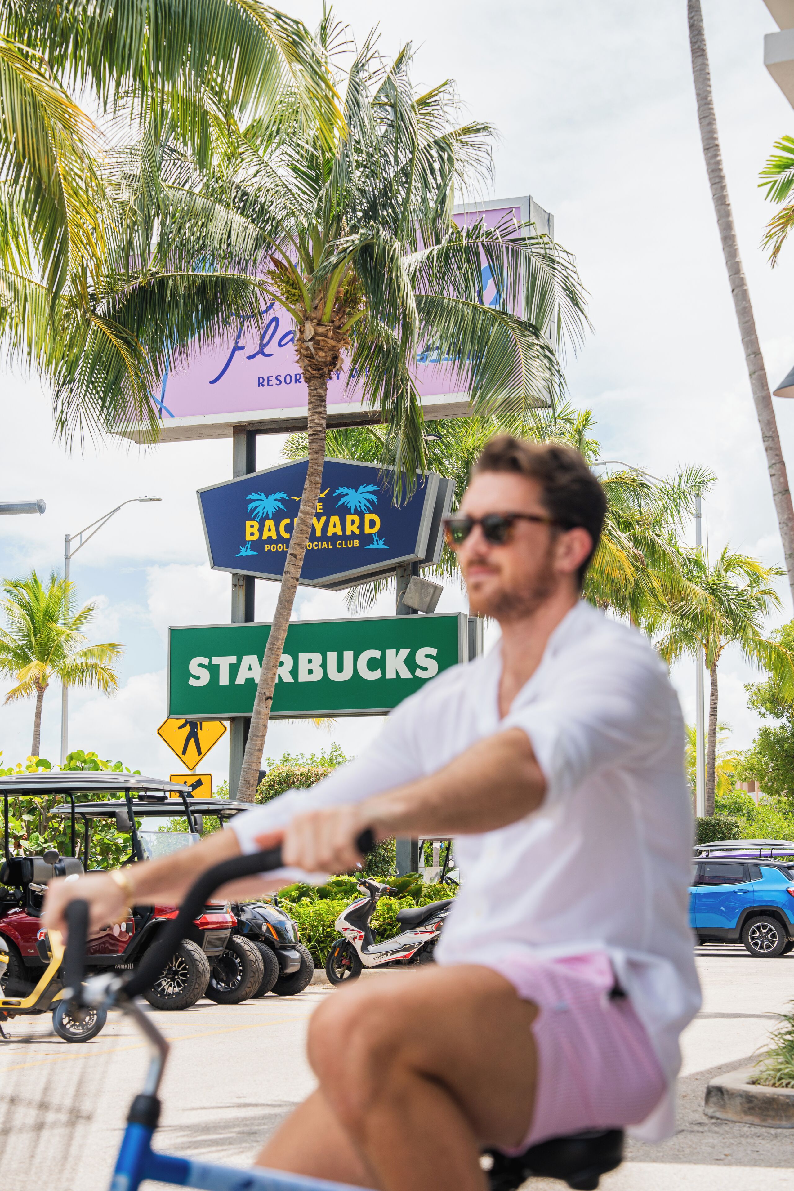 A person riding a bicycle on a street with palm trees and signs for a pool and Starbucks.
