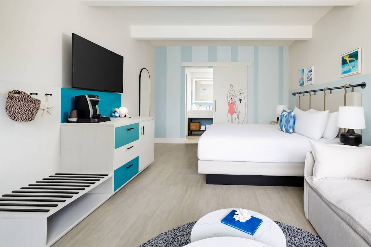 A modern hotel room with a bed, a sofa, a TV, a desk, and a bathroom visible through an open door.