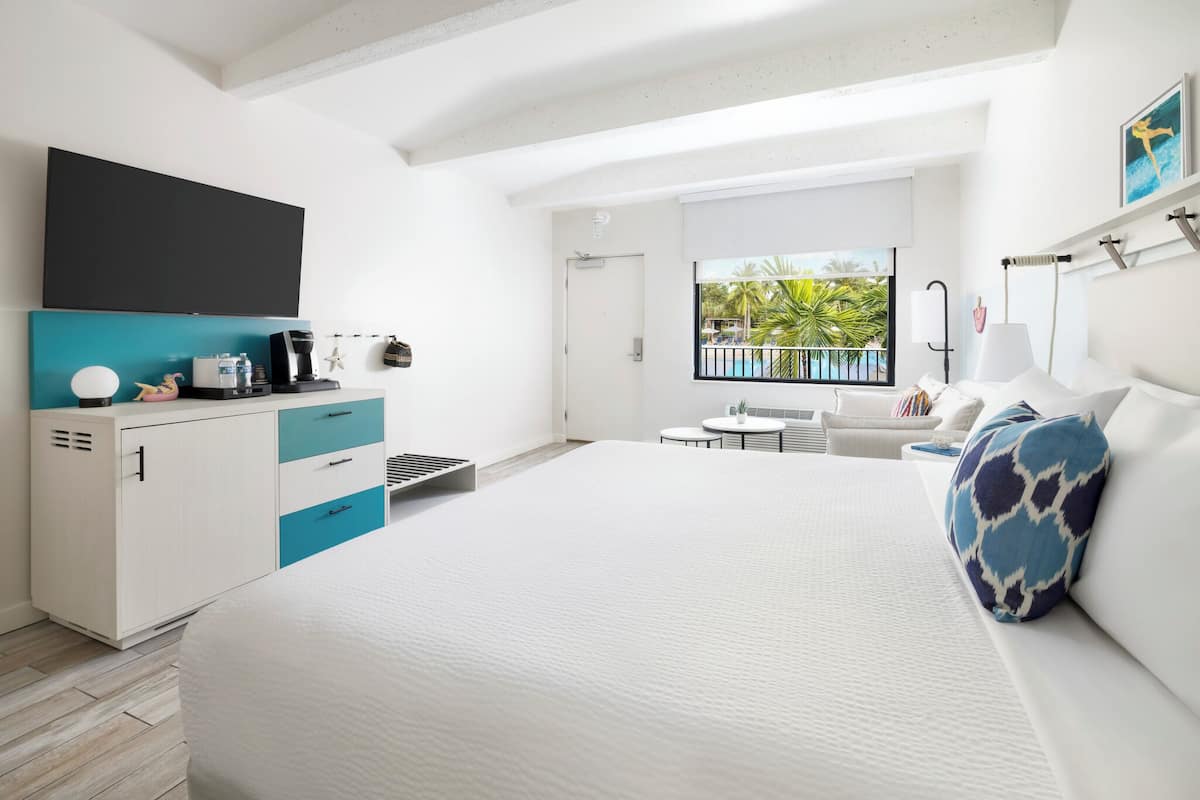 A modern hotel room with a large bed, a small desk with a computer, a window with a view of palm trees, and a balcony with a pool.