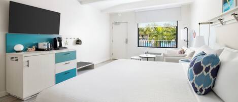 A modern hotel room with a large bed, a small desk with a computer, a window with a view of palm trees, and a balcony with a pool.