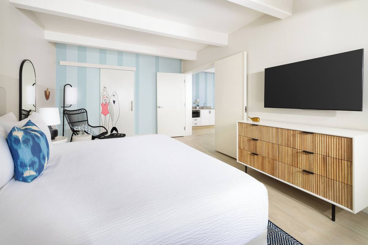 A modern hotel room with a large bed, a flat-screen TV, a wooden cabinet, and a view into the kitchen area.
