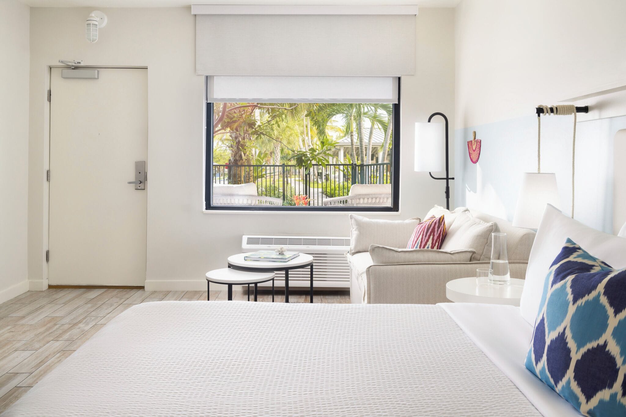 A hotel room with a large bed, a small round table, a sofa, a window with a view of palm trees, and a door.