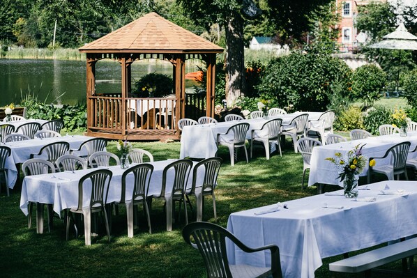 An outdoor dining area with tables and chairs arranged around a gazebo by a lake.