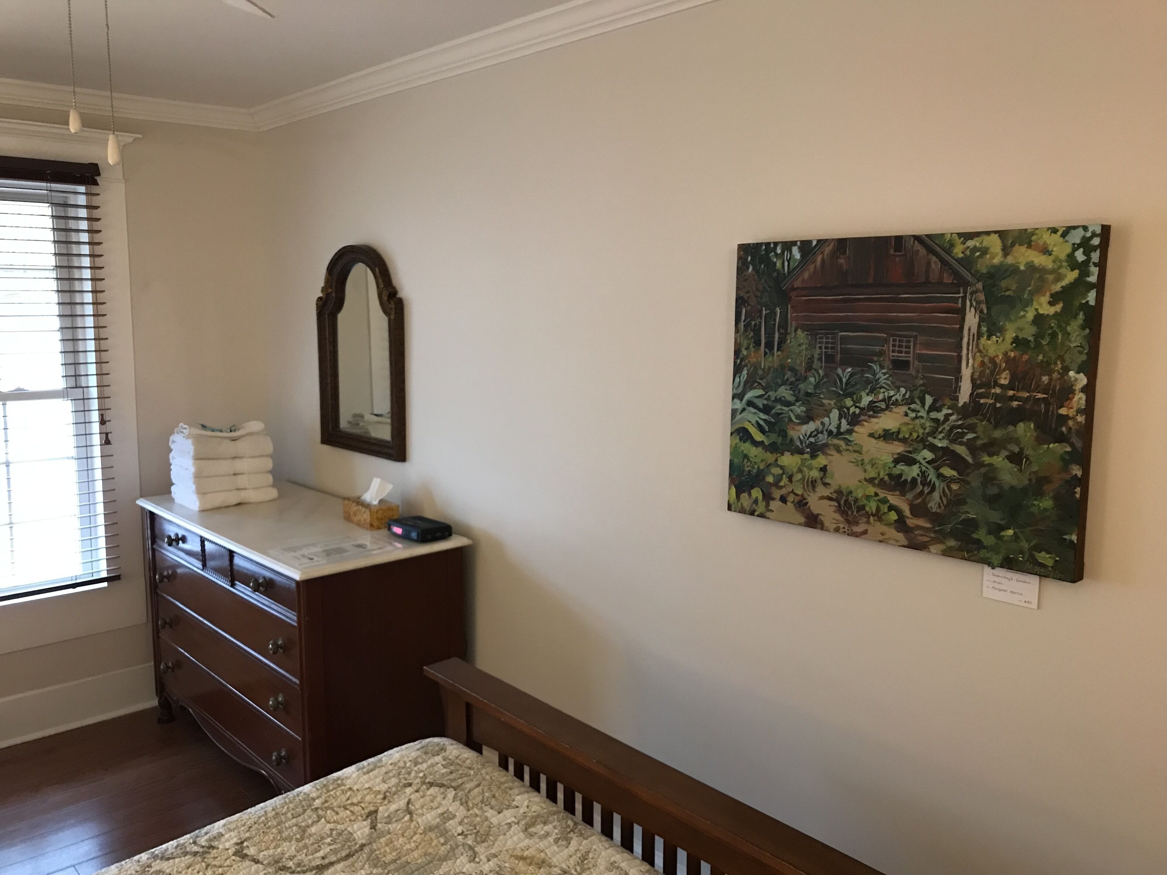 A bedroom with a wooden dresser, a bed with a headboard, a mirror, and a painting on the wall.
