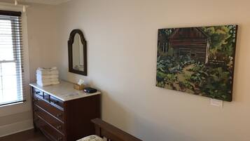 A bedroom with a wooden dresser, a bed with a headboard, a mirror, and a painting on the wall.