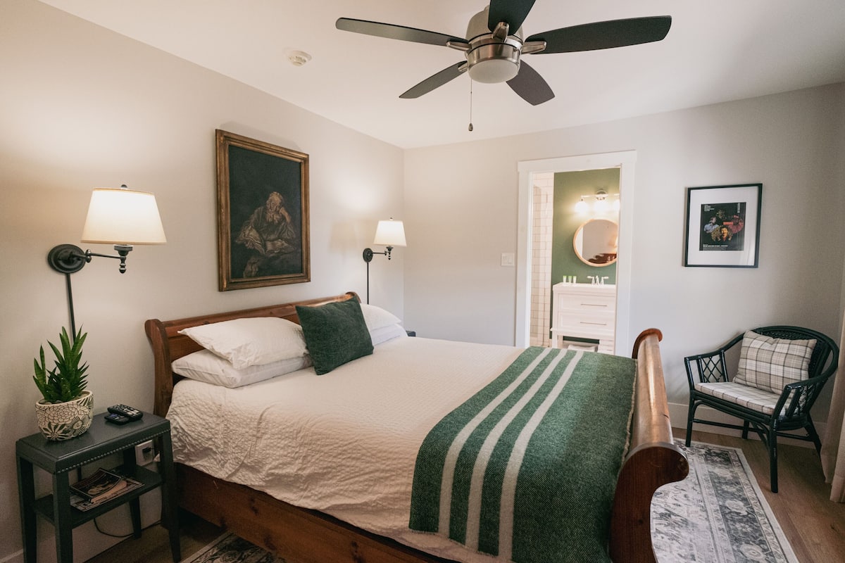 Une chambre à coucher comprenant un lit, des lampes de chevet, un ventilateur de plafond, une chaise et une porte donnant sur une salle de bain.