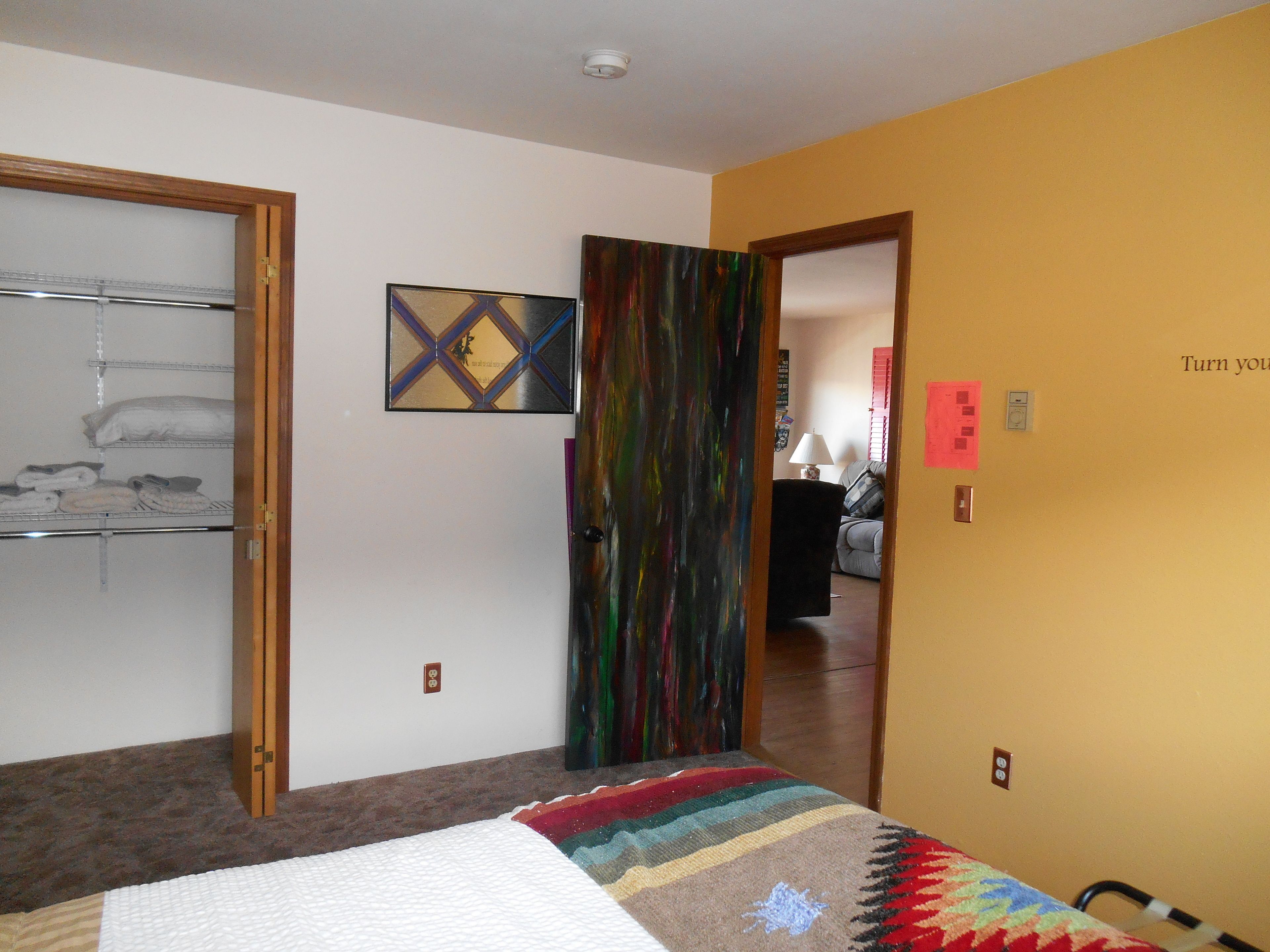 A room with a bed, a door leading to another room, a shelf with folded items, and a painting on the wall.