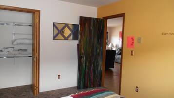A room with a bed, a door leading to another room, a shelf with folded items, and a painting on the wall.