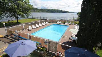 Une piscine entourée d’une clôture, dotée de chaises longues et de parasols, et offrant une vue sur un lac et des arbres.