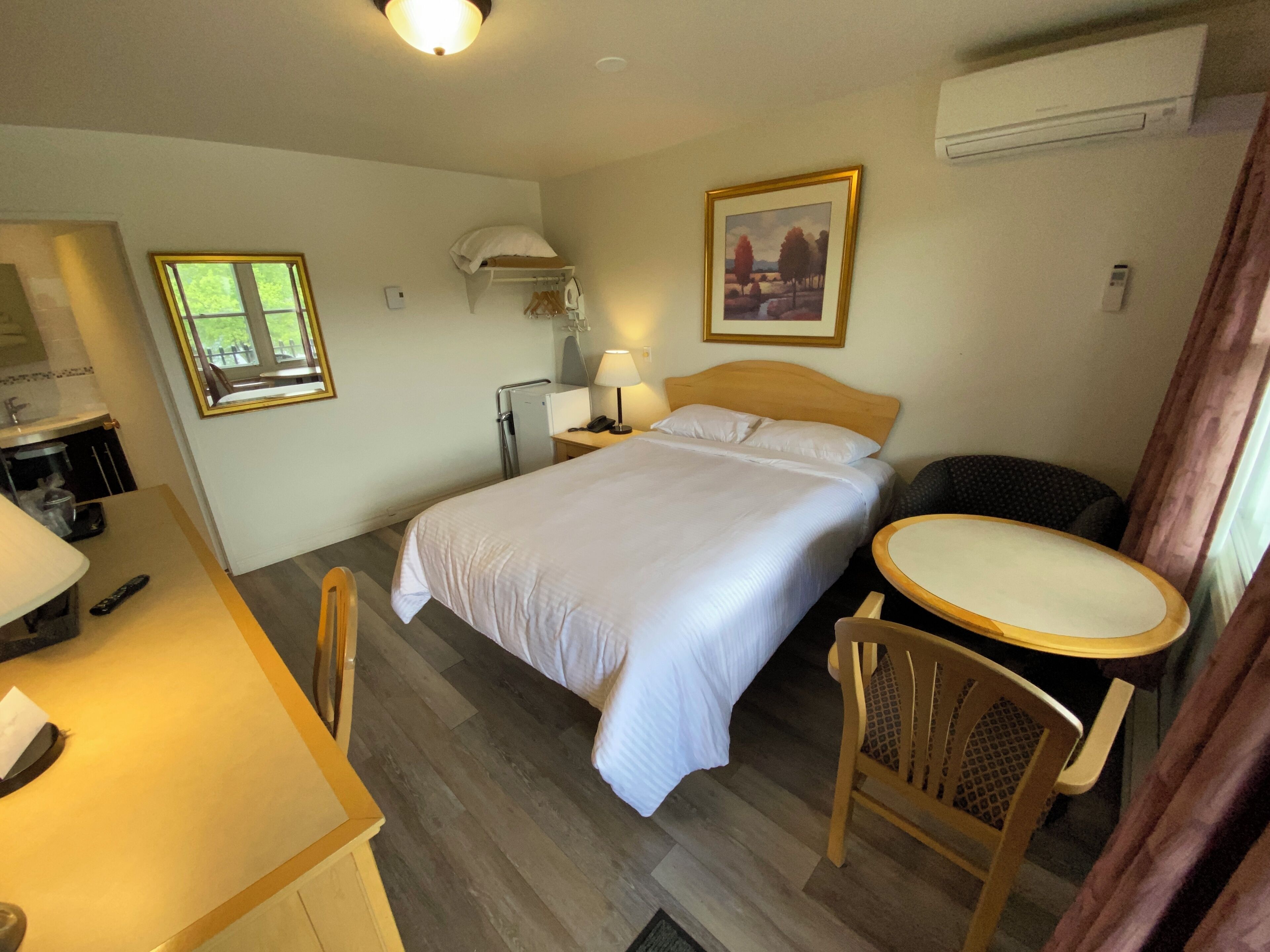 A hotel room with a bed, a desk with a chair, a small table, a lamp, a painting on the wall, and an air conditioning unit.