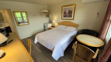 A hotel room with a bed, a desk with a chair, a small table, a lamp, a painting on the wall, and an air conditioning unit.