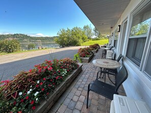 A patio with outdoor seating, a view of a lake, and a building with a covered entrance. - Motel Panoramique (Saguenay)