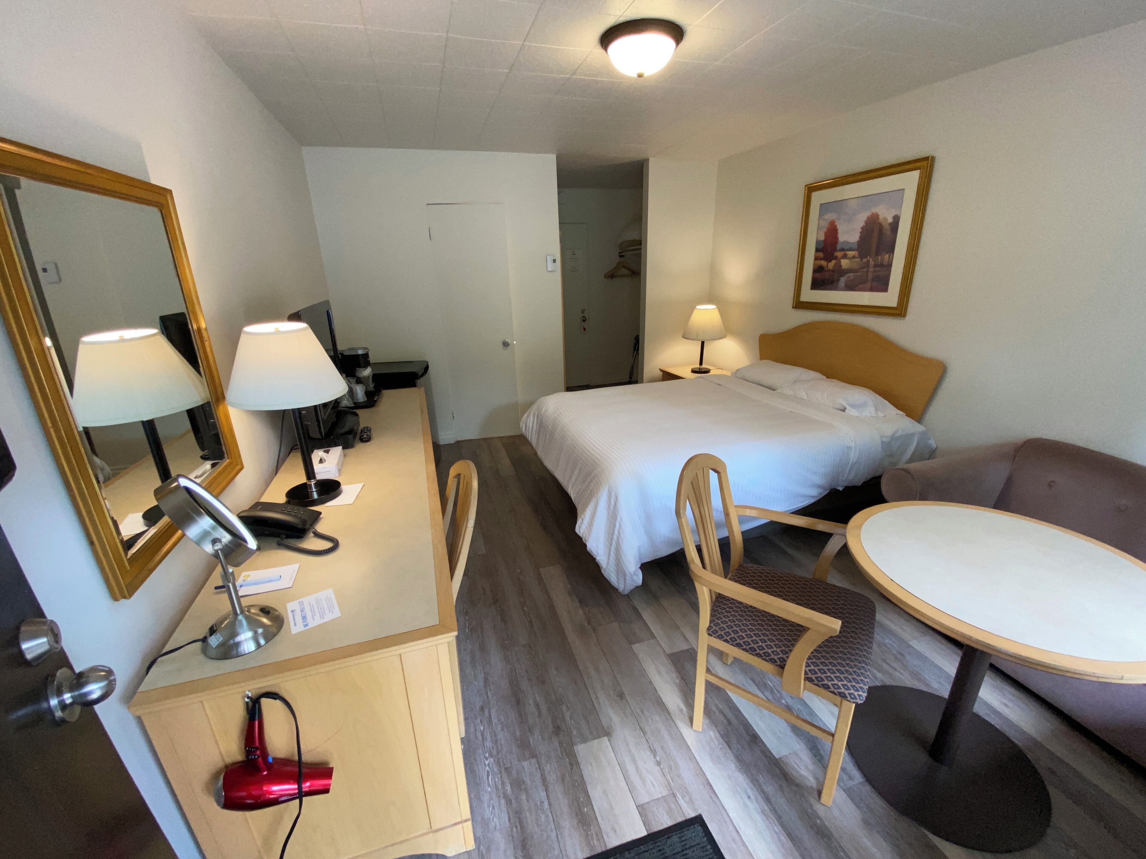 Habitación de hotel con cama, escritorio con lámpara, silla, mesita pequeña, secador de pelo rojo y un cuadro en la pared.