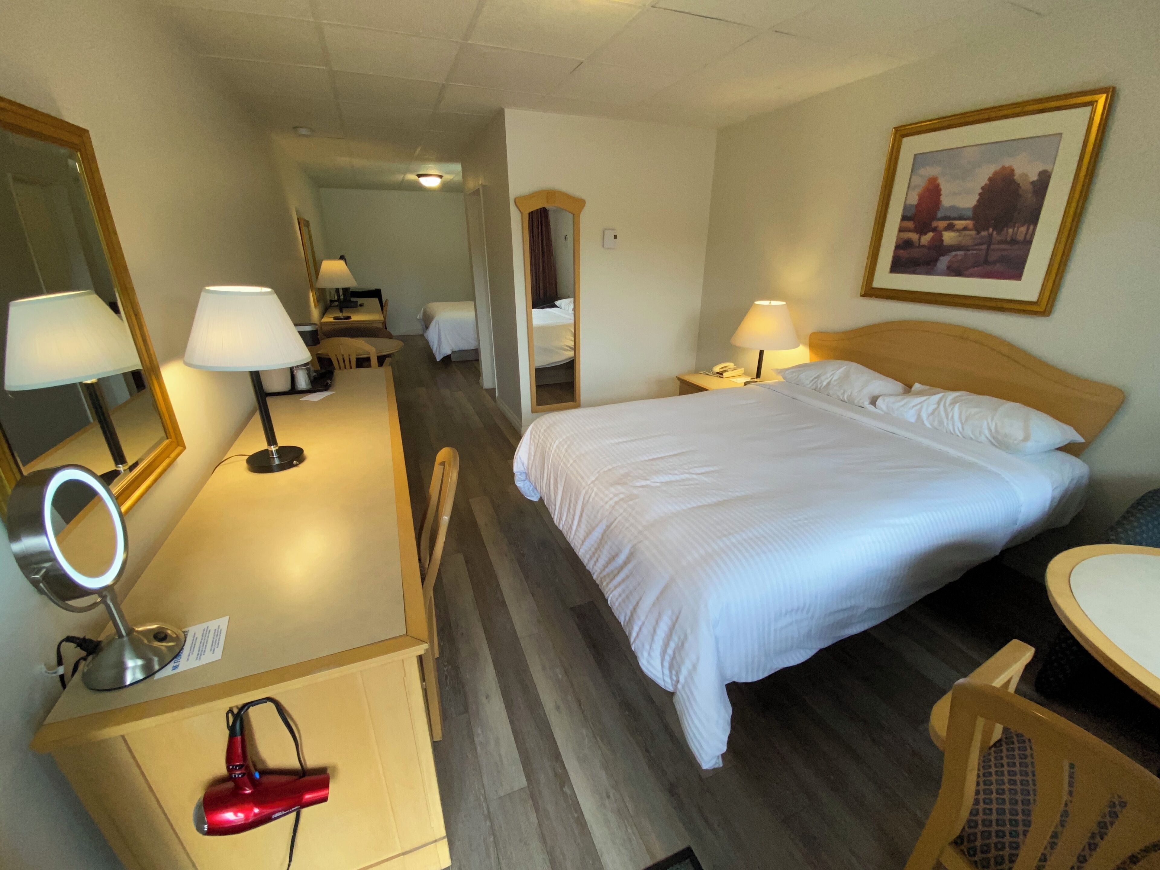 Ein Hotelzimmer mit Bett, Schreibtisch, Stuhl und Spiegel. Auf dem Schreibtisch steht eine Lampe und auf dem Stuhl ein roter Föhn.