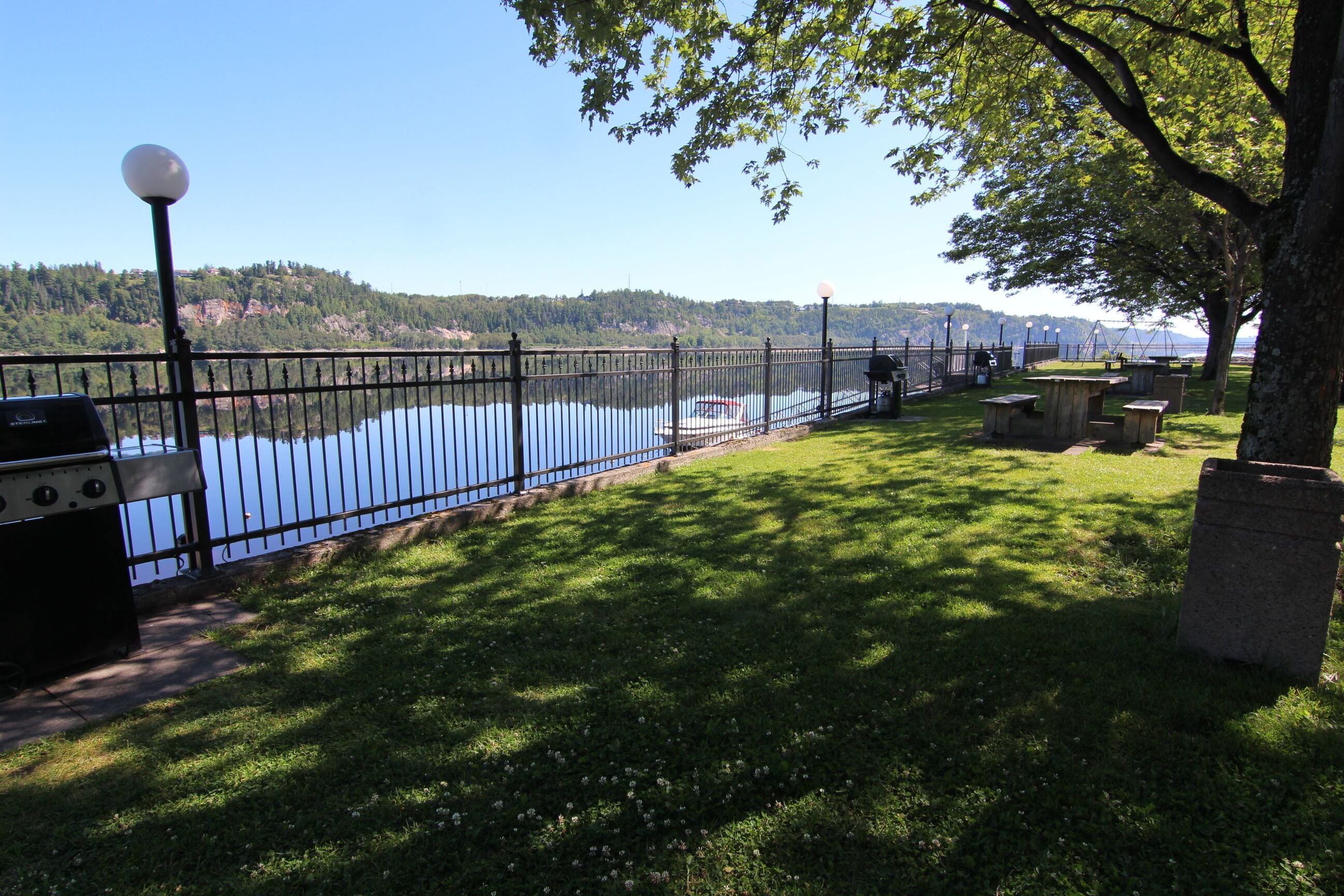 A lakeside park with a metal fence, picnic tables, and a barbecue grill.