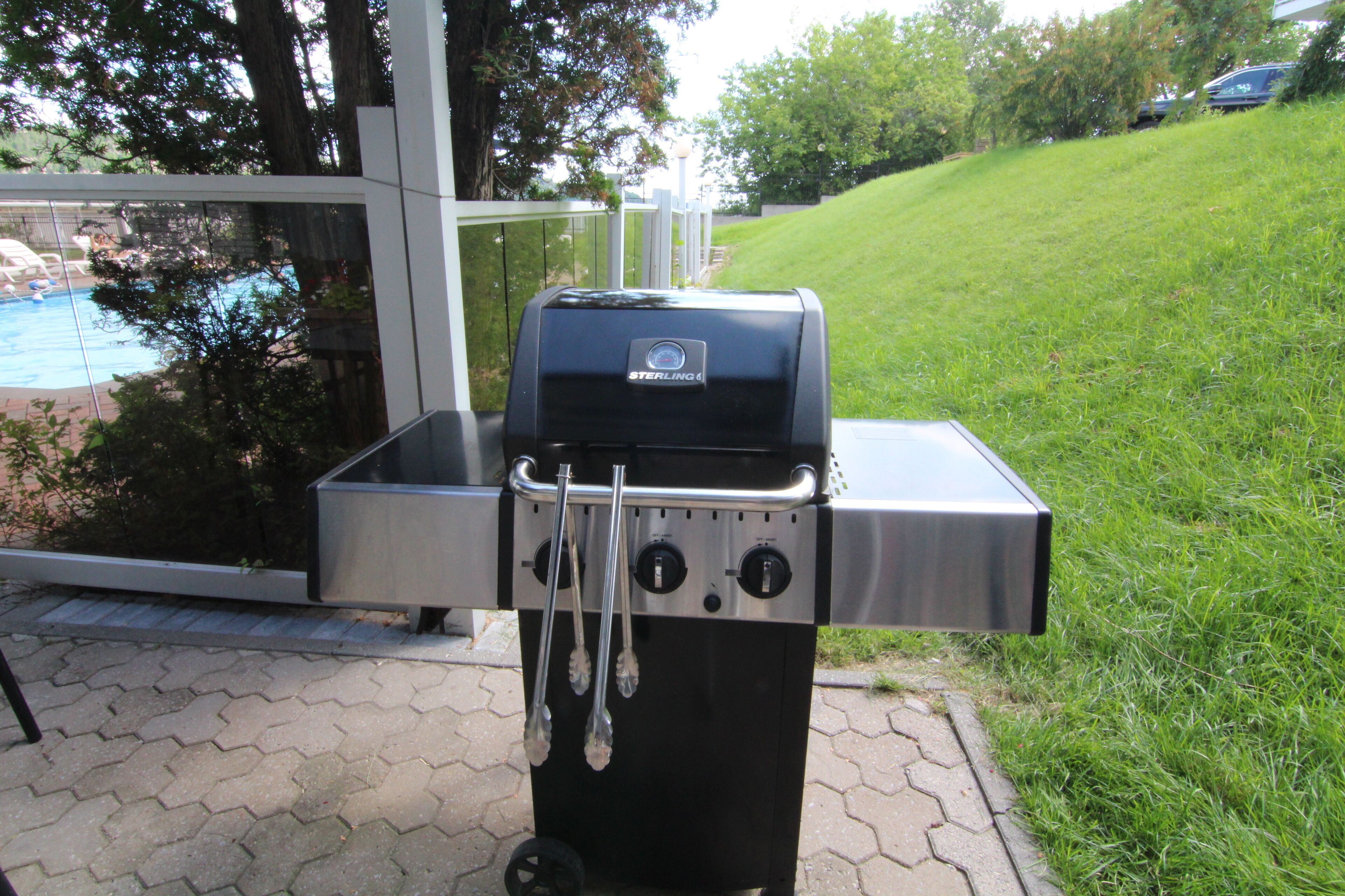 A stainless steel grill with two side burners and a lid, placed on a paved patio with a pool visible in the background.