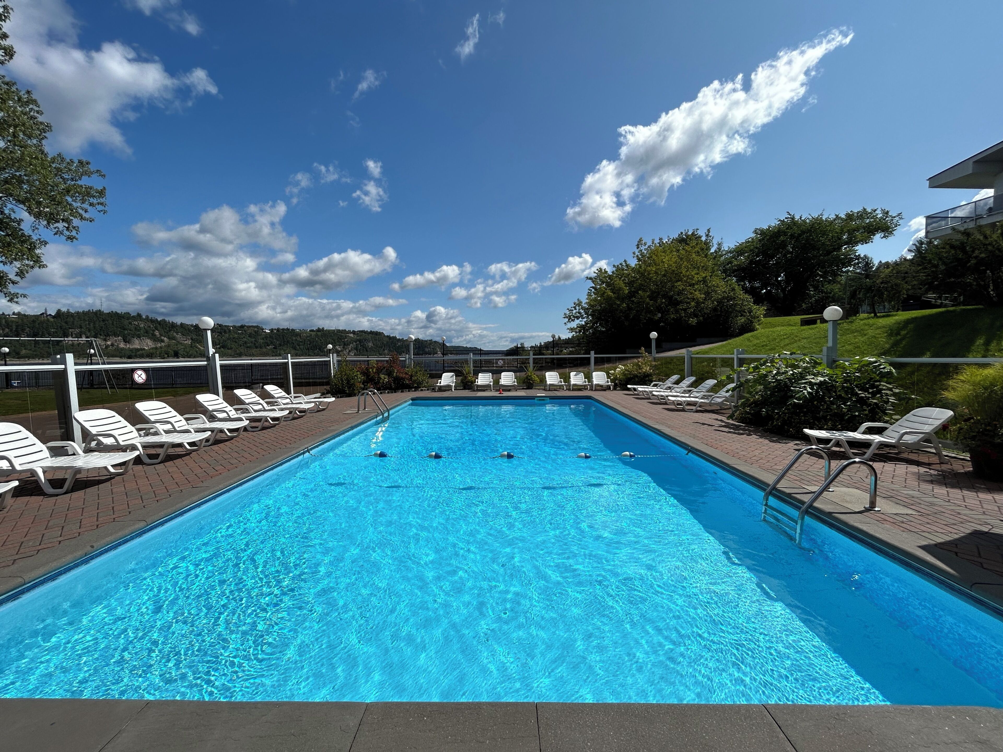 A rectangular swimming pool with clear blue water, surrounded by white lounge chairs and a brick walkway.