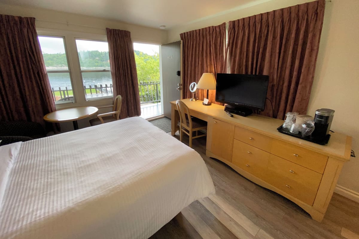 A hotel room with a bed, a desk with a TV, a chair, a small table, and a view of a body of water through large windows.