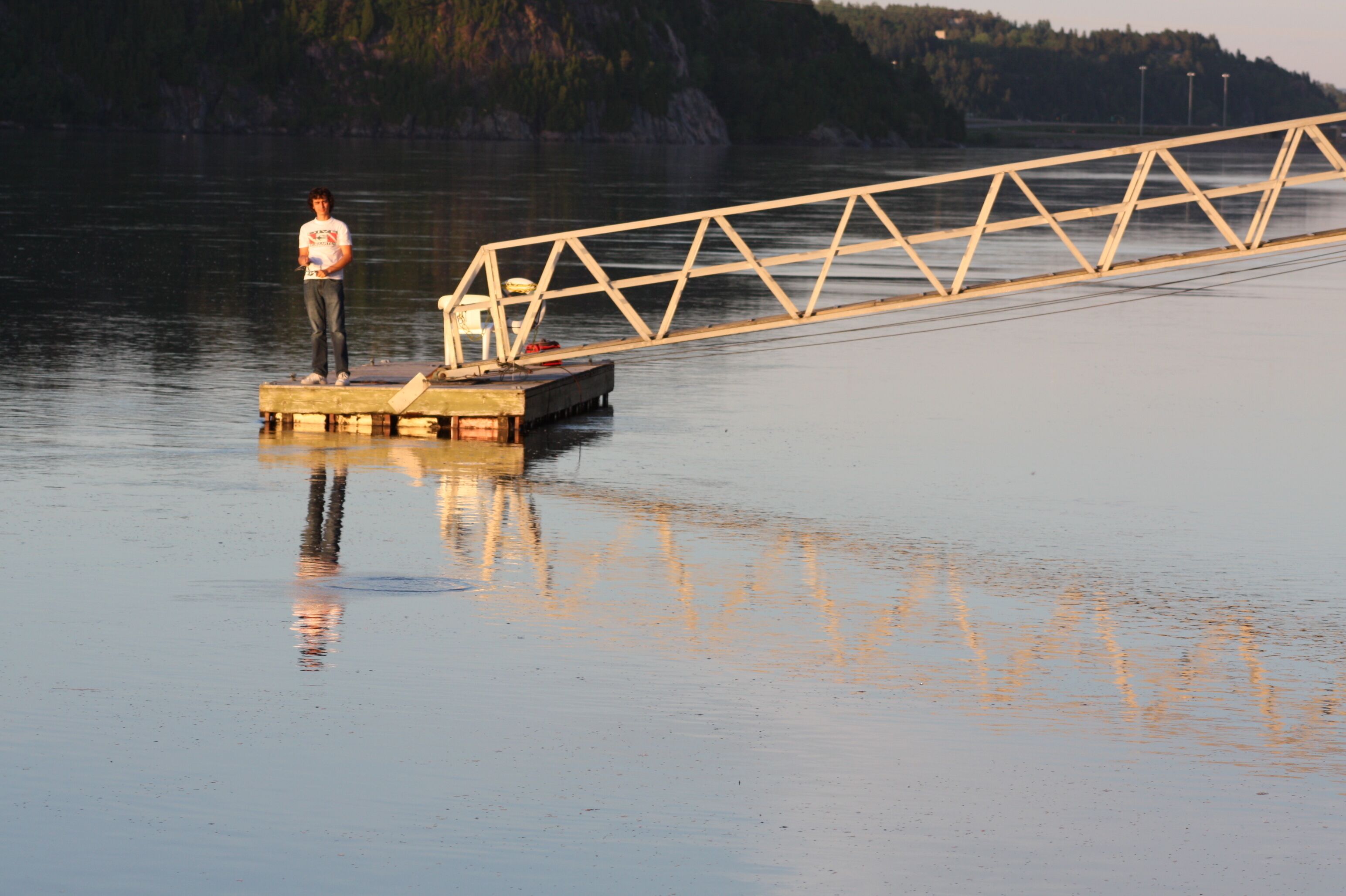 Une personne se tient sur une jetée en bois qui s’avance sur une étendue d’eau calme, avec une structure de pont métallique en arrière-plan.