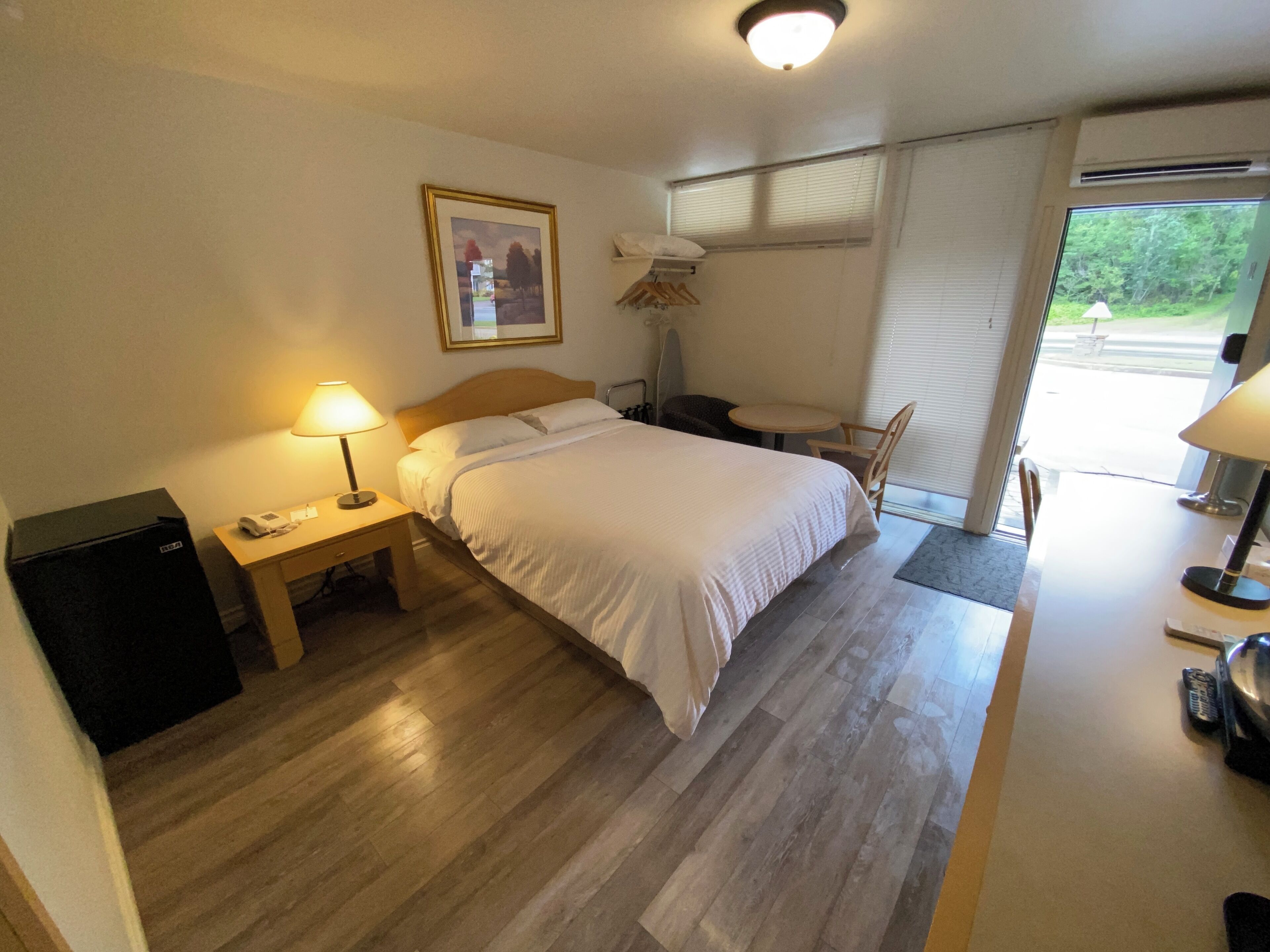 A hotel room with a bed, bedside table with a lamp, a small desk with a chair, a window with blinds, and a view of greenery outside.